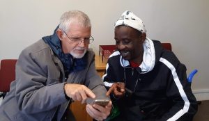A grey-haired man shows a younger man how to access applications on a smart phone that help communication