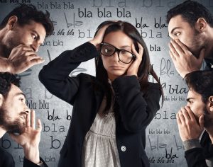 Image showing a young woman with long dark hair and wearing glasses with a black frame and a black jacket, with her hands in her hair, while faces of men around her are speaking. There are words floa ting in the space around her.